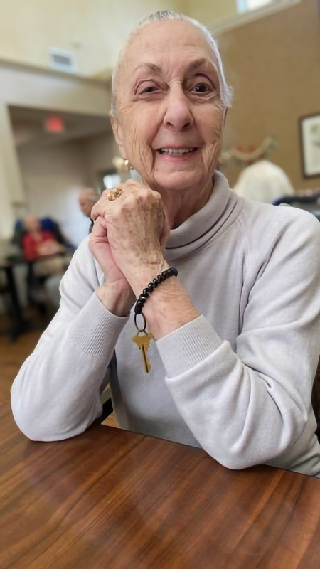 Joan is wearing a long-sleeved white turtleneck and sitting at a table with folded hands in The Parkway Senior Living.