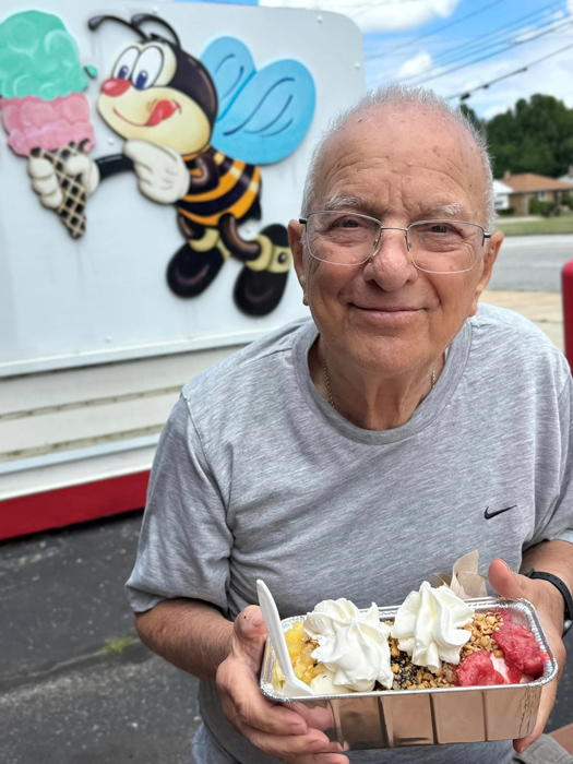 Senior resident smiles while holding a banana split topped with whipped cream, standing in front of a colorful mural of a bee holding an ice cream cone on a sunny day.