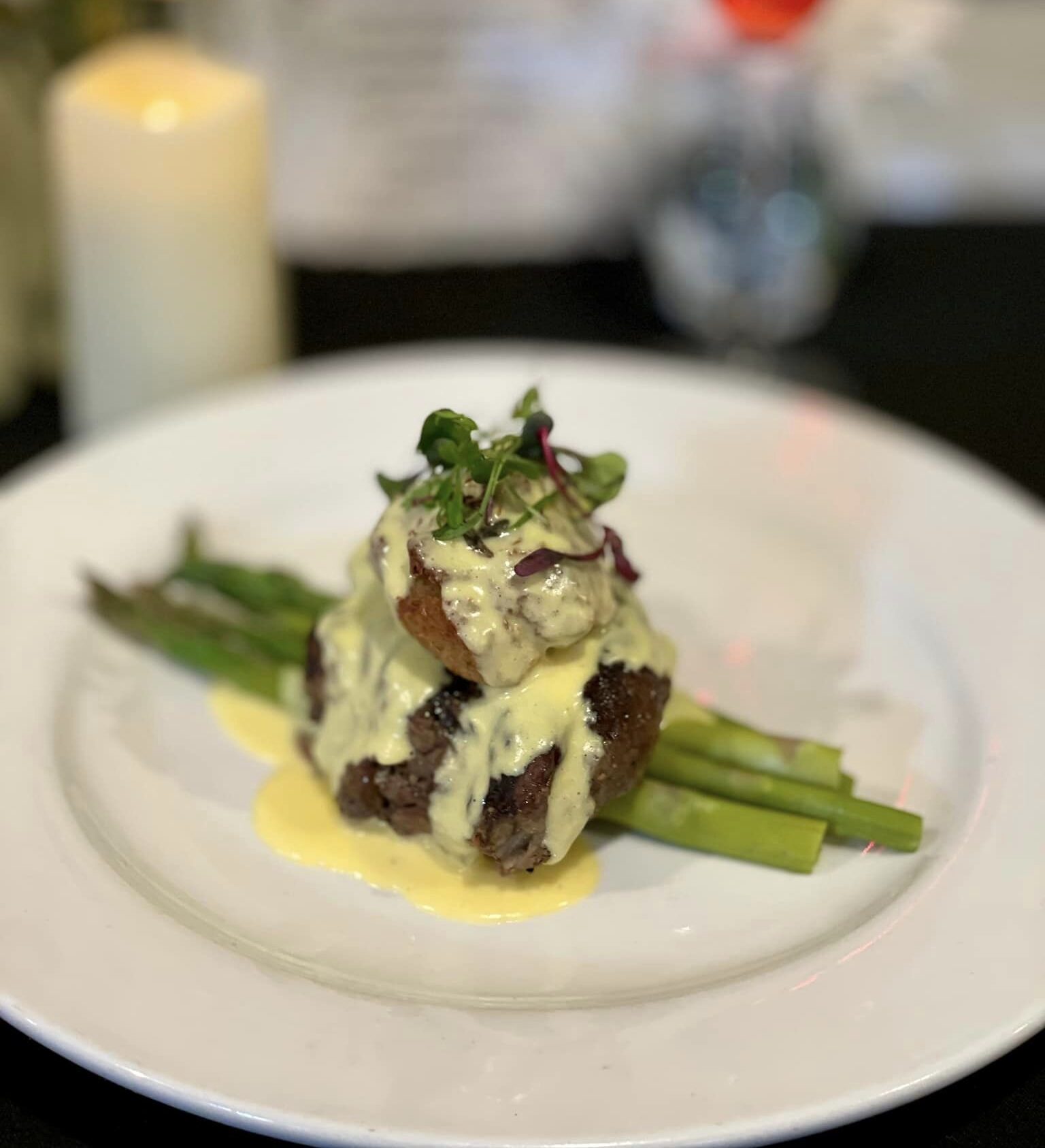 Plated gourmet steak with asparagus, topped with creamy hollandaise sauce and garnished with microgreens, set against a blurred background of a candle, flowers, and drink.Plated gourmet steak with asparagus, topped with creamy hollandaise sauce and garnished with microgreens, set against a blurred background of a candle, flowers, and drink.