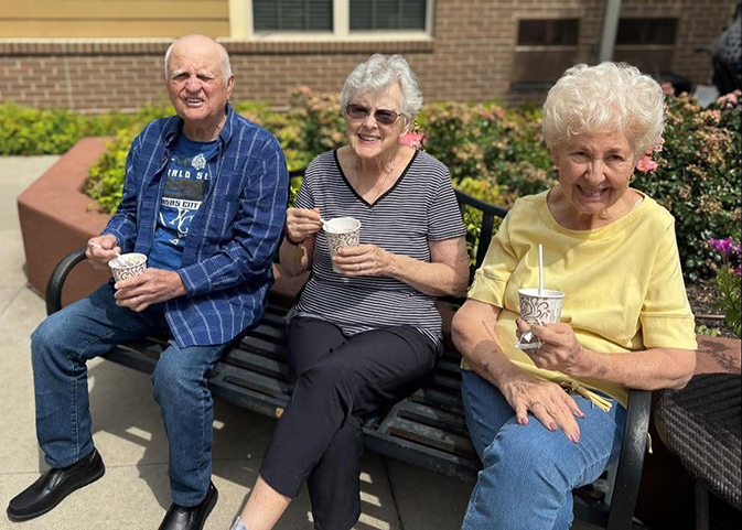 Three senior residents enjoy ice cream outdoors on a sunny day, smiling while seated on a bench near flowers.