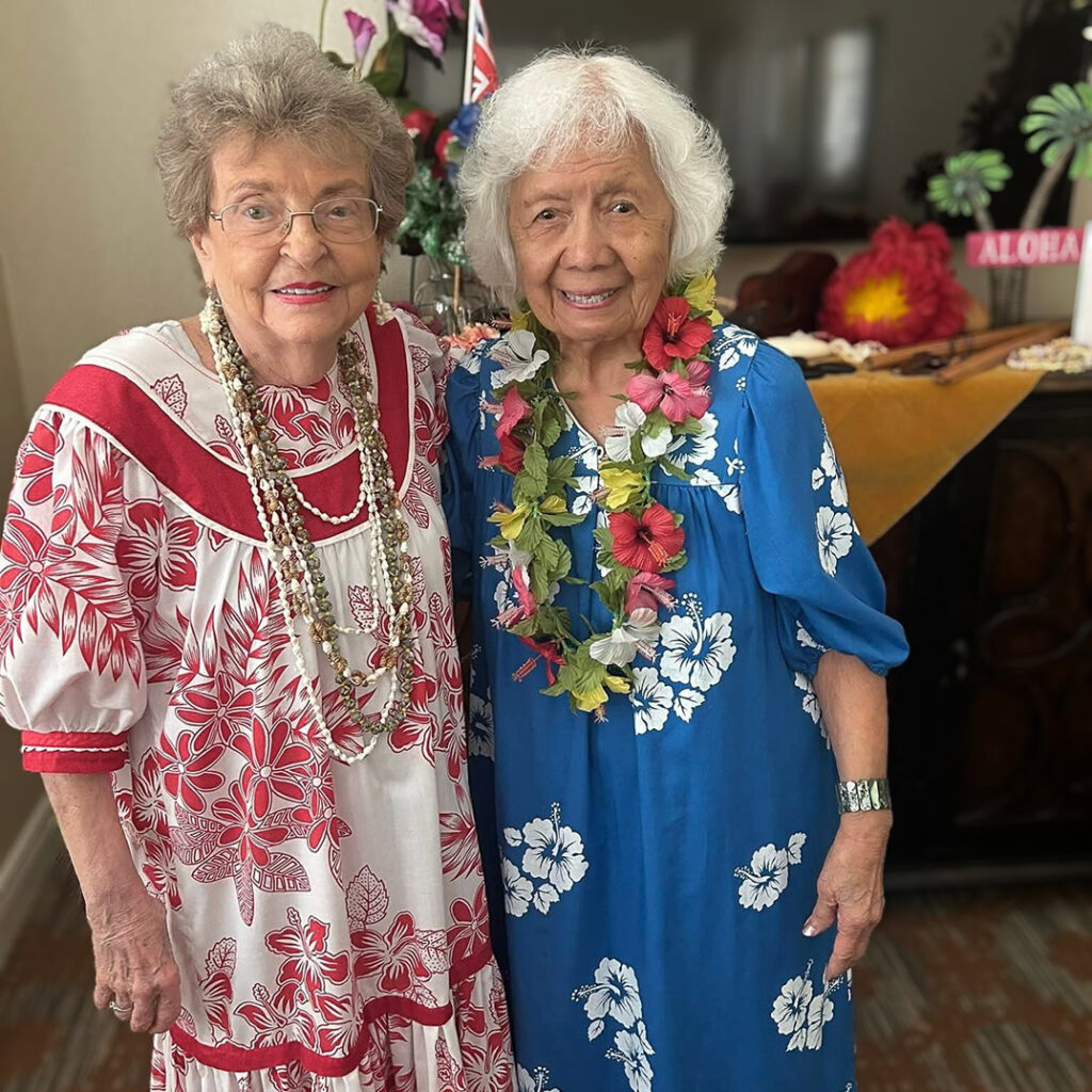 Two senior women dressed in Hawaiian floral attire with leis smile together during a themed luau celebration.
