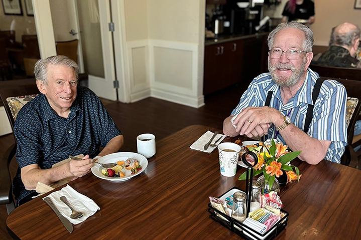 Two senior men share a meal and coffee at a dining table, smiling warmly during a casual breakfast gathering at their senior living community.