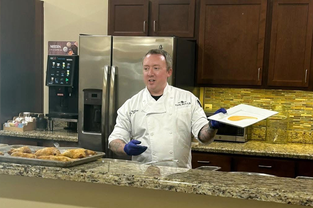 Chef at The Parkway Senior Living demonstrates a dish, holding a plated sauce while baked pastries rest on the counter during a community culinary presentation.