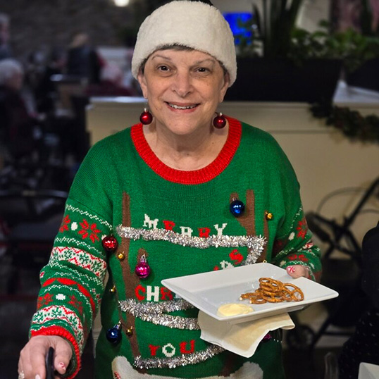 A woman in a Christmas sweater and Santa hat smiles while helping herself to festive food during a gathering.