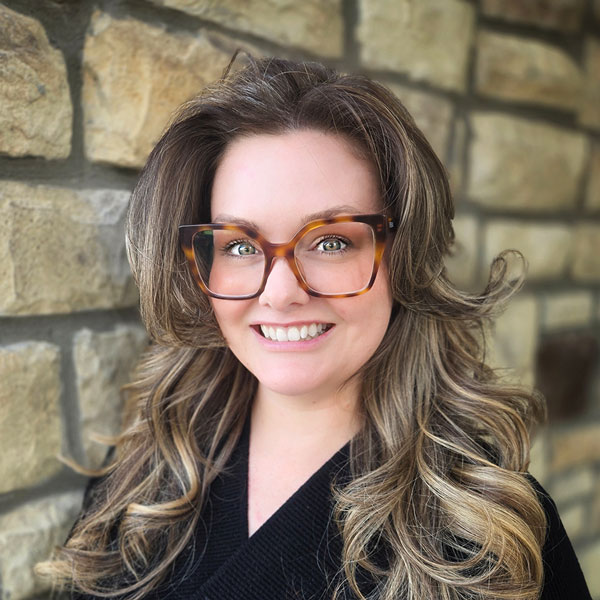 Emily Hornback, Wellness Director at The Parkway Senior Living, smiling in front of a tan stone wall while wearing large tortoiseshell glasses and a black top.