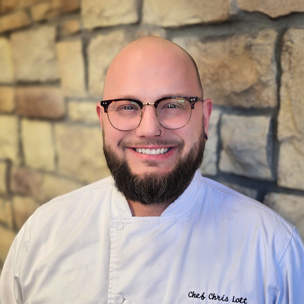 Chris Lott, Culinary Director at The Parkway Senior Living, smiling in a white chef coat while standing indoors against a stone wall background.
