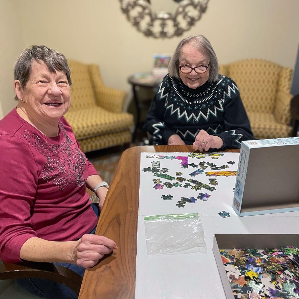 Two senior women smile while working on a puzzle together.