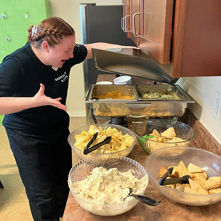 A team member gestures excitedly, opening a large dish of food on a prepared meal buffet for the team.