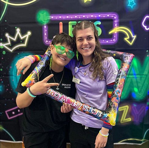 Two team members at The Parkway Senior Living smile, sporting brightly colored props beside a neon background during a celebration.