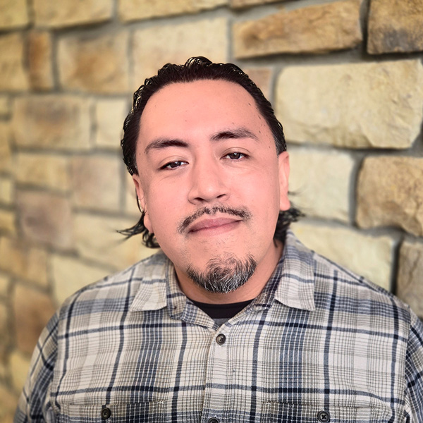 Ernesto Perez, Plant Operations Director at The Parkway Senior Living, smiling in a professional headshot, wearing a plaid button-down shirt, with a stone wall background.