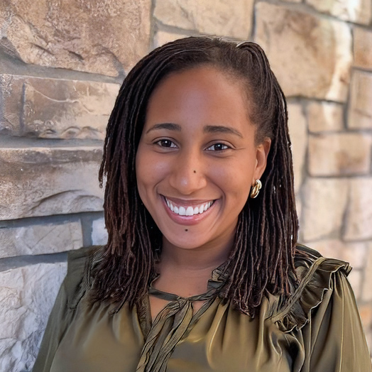 Asia Latta, Business Office Director at The Parkway Senior Living, smiling in a professional headshot with shoulder-length locs, gold earrings, an olive green blouse, and a light stone wall background.