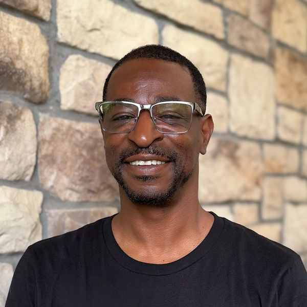 Eric Kerns, Plant Operations Director at The Parkway Senior Living, smiling in a professional headshot with short hair, glasses, a trimmed goatee, a black shirt, and a stone wall background.