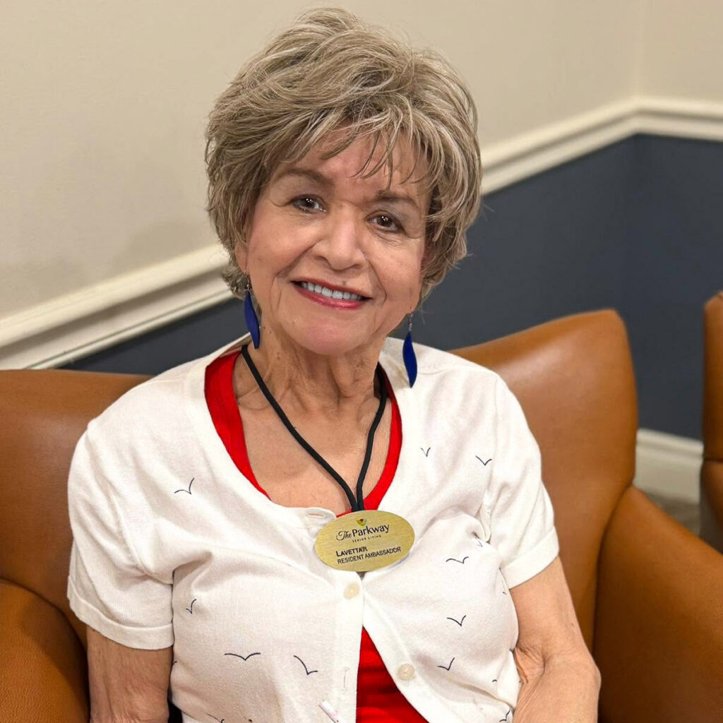 A woman on the Resident Ambassador team at The Parkway Senior Living smiles brightly wearing a gold ambassador badge.