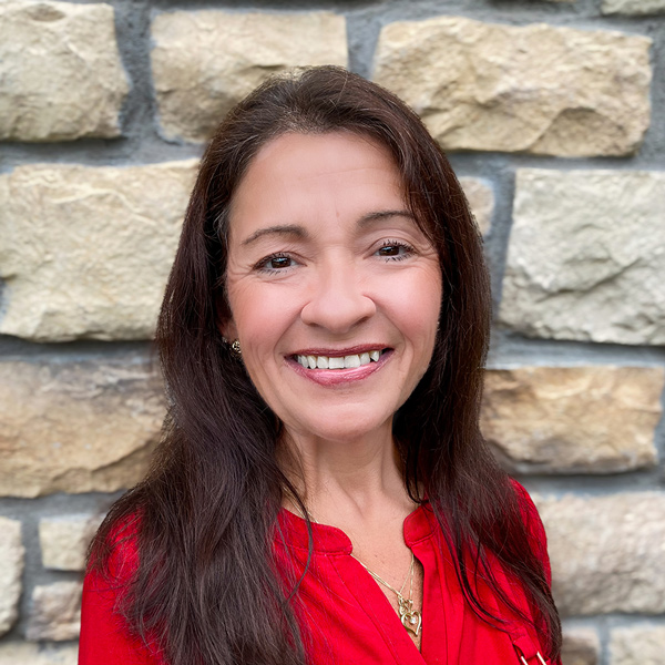 Andrea Groh, Senior Living Coordinator at The Parkway Senior Living, smiling in a professional headshot with long dark hair, a red blouse, and a light stone wall background.