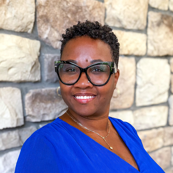 Marilyn Dove, Business Office Director at The Parkway Senior Living, smiling in a professional headshot with short curly hair, oversized black glasses, a royal blue blouse, and a light stone wall background.